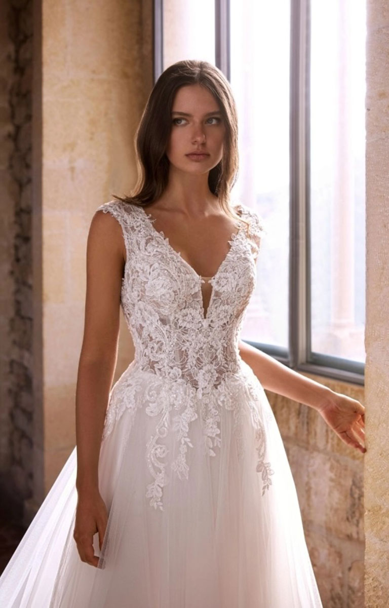 A woman wearing Gemmah by Étoile Bridal - Beaded Lace A-Line Wedding Dress with V-Neckline and Royal Train with a deep V-neckline and sheer embroidered bodice, standing indoors near a window with warm stone walls.