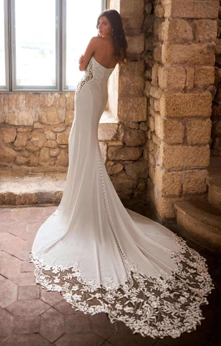A woman wearing Ginessa by Étoile Bridal - Beaded Lace Fit-and-Flare Wedding Dress with Strapless Scoop Neckline with a long lace-trimmed train, posed indoors against a stone wall and window.