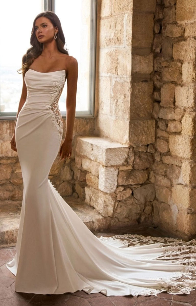 A woman models Ginessa by Étoile Bridal - Beaded Lace Fit-and-Flare Wedding Dress with Strapless Scoop Neckline with a ruched bodice, lace side detailing, and a long flowing train in an indoor stone setting.