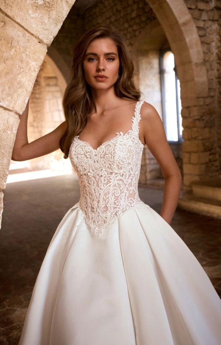 A woman wearing Glorielle by Étoile Bridal - Satin Ballgown Wedding Dress with Sweetheart Neckline and Royal Train stands in a stone archway corridor. The dress has a fitted lace bodice with thin straps and a full satin skirt.