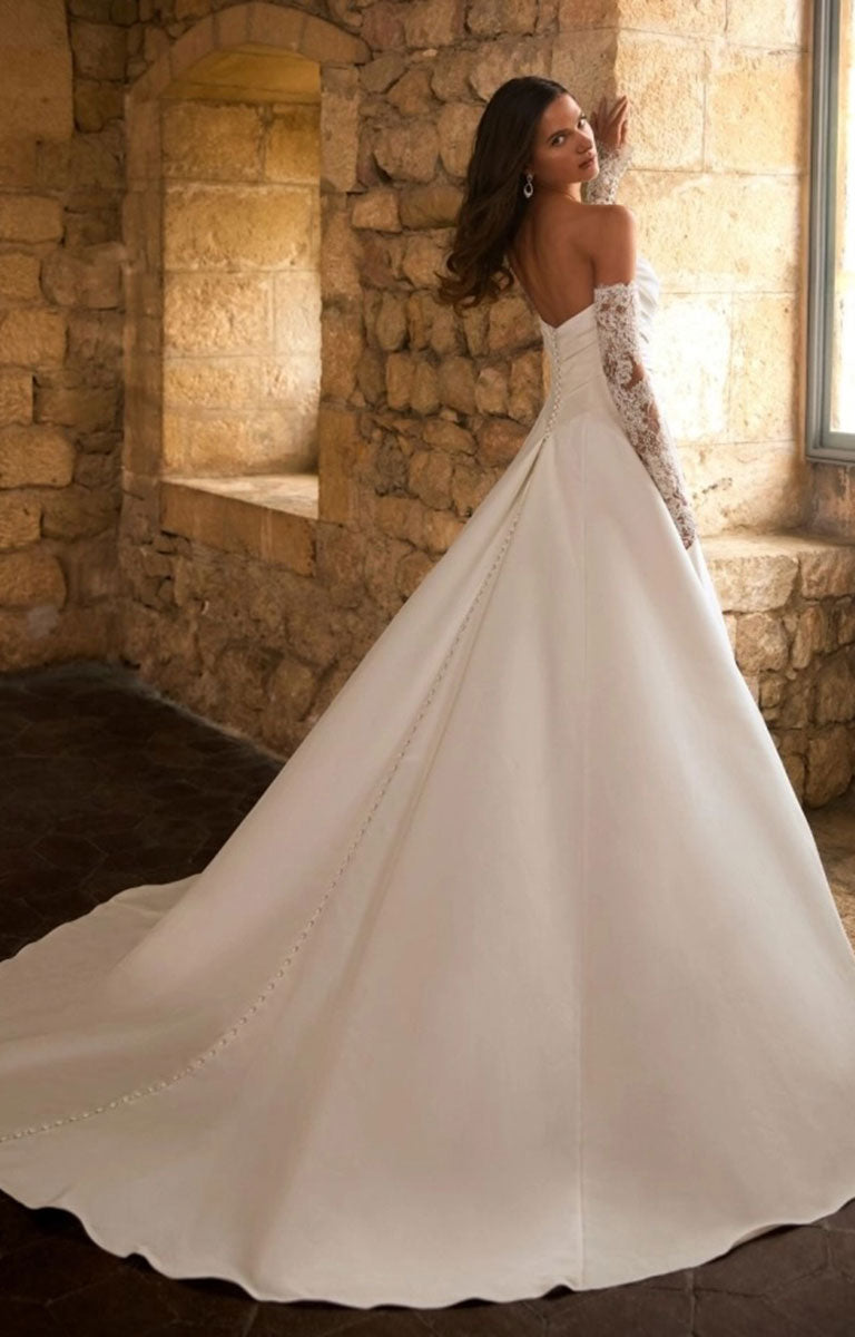 A woman wearing Gracelle by Étoile Bridal - Satin Ballgown Wedding Dress with Detachable Lace Sleeves and Royal Train with lace sleeves, standing in a stone interior with warm sunlight streaming in from a window.