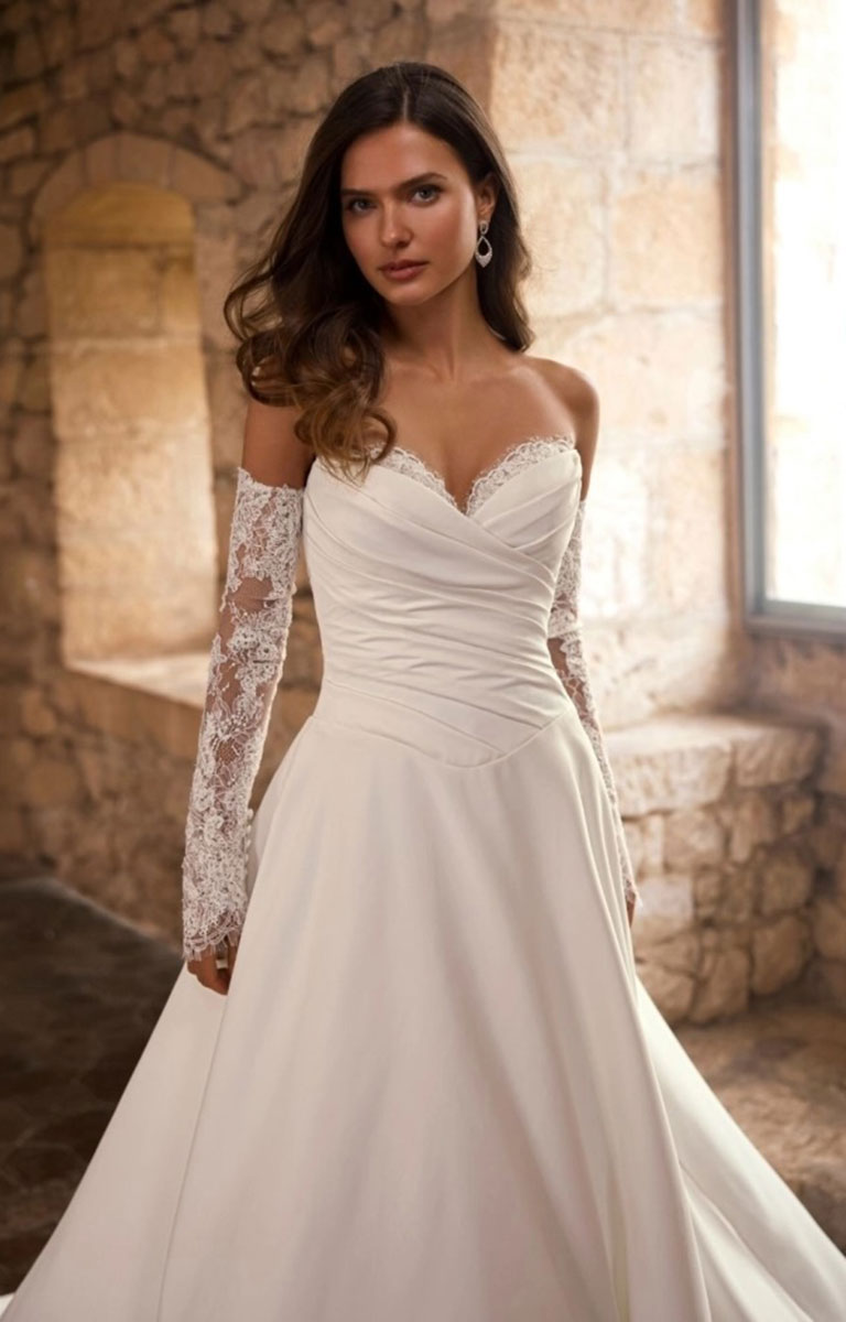 A woman wearing Gracelle by Étoile Bridal - Satin Ballgown Wedding Dress with Detachable Lace Sleeves and Royal Train with lace sleeves, standing in a stone interior with warm sunlight streaming in from a window.
