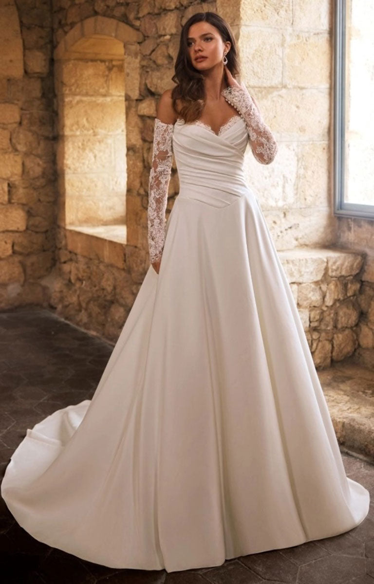 A woman wearing Gracelle by Étoile Bridal - Satin Ballgown Wedding Dress with Detachable Lace Sleeves and Royal Train with lace sleeves, standing in a stone interior with warm sunlight streaming in from a window.