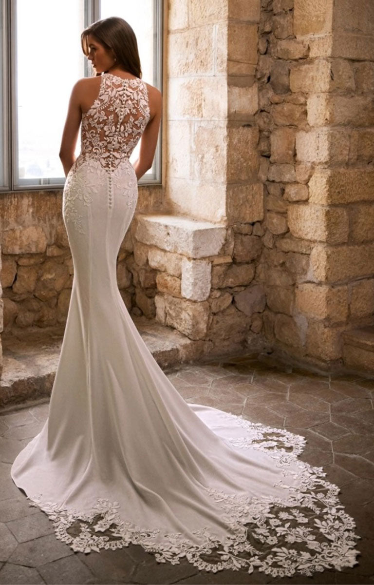 Woman wearing Gracy by Étoile Bridal - Mermaid Wedding Dress with Halter Neckline, Basque Waist and Botanical Lace standing facing a window in a warm honeyed colour stone wall with soft lighting 