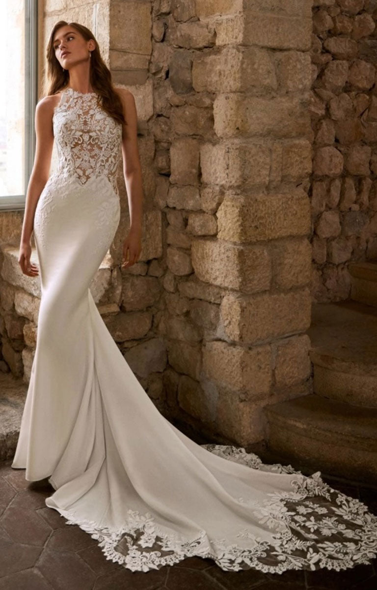 Woman wearing Gracy by Étoile Bridal - Mermaid Wedding Dress with Halter Neckline, Basque Waist and Botanical Lace standing against a window in a warm honeyed colour stone wall with soft lighting 