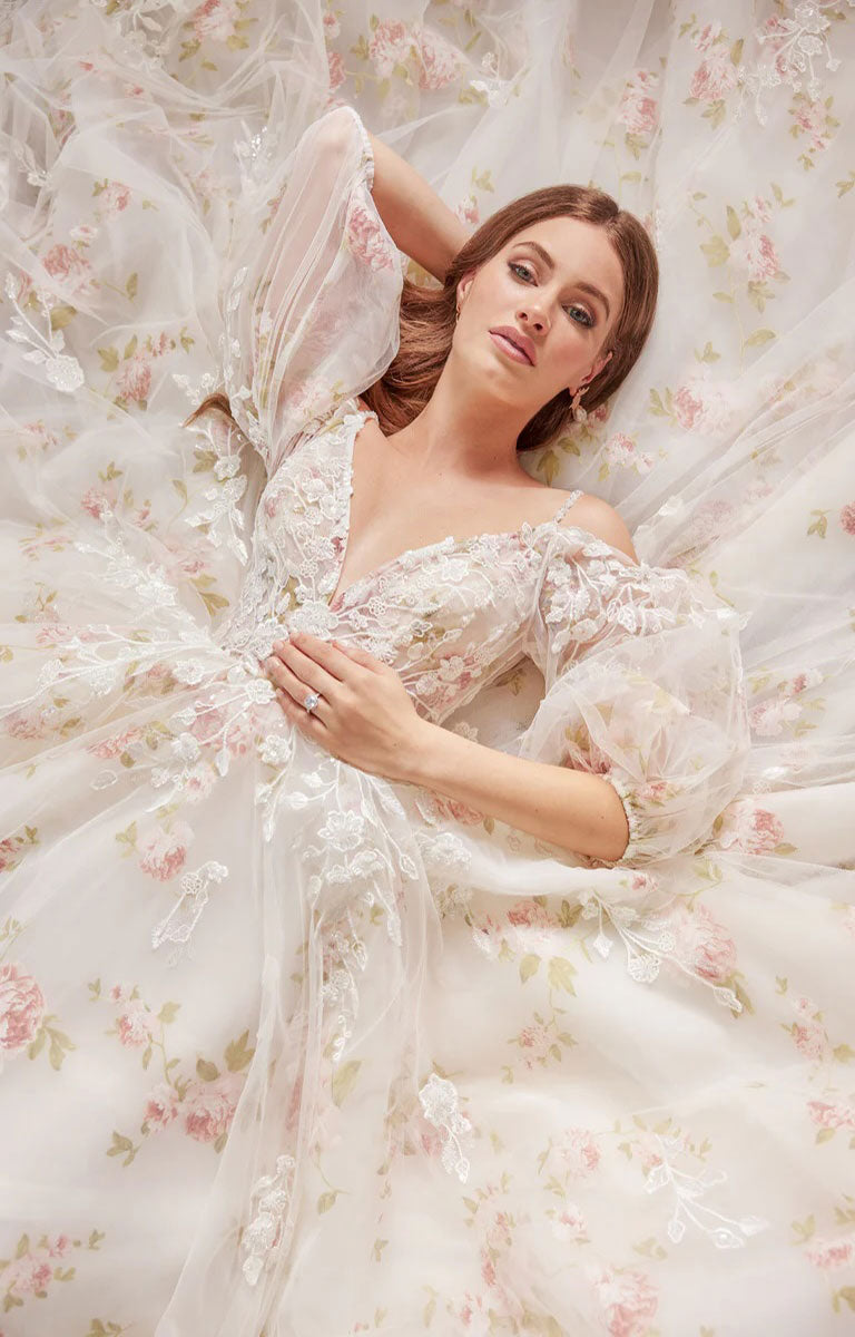 Woman in Elora by Josephine Scott - Romantic Rose Print Ballgown Wedding Dress with Lace Appliqué and Voluminous Bell Sleeves lying on a floral background