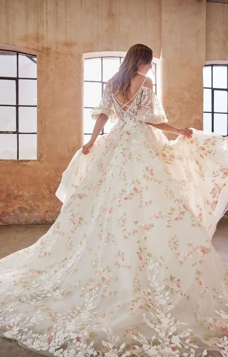 Woman in Elora by Josephine Scott - Romantic Rose Print Ballgown Wedding Dress with Lace Appliqué and Voluminous Bell Sleeves standing in a room with large windows.