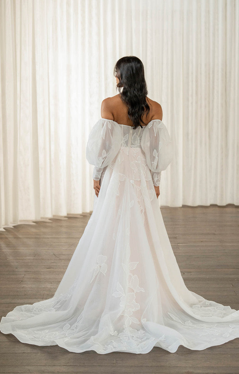 Woman wearing Tessa by Madi Lane, a white wedding dress with sheer sleeves in front of white curtains.