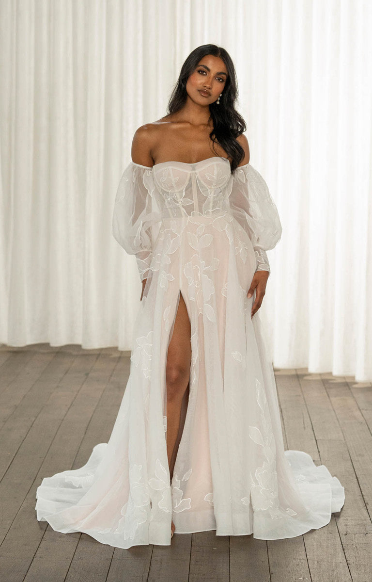 Woman wearing Tessa by Madi Lane, a white lace wedding dress with sheer sleeves in a softly lit room.