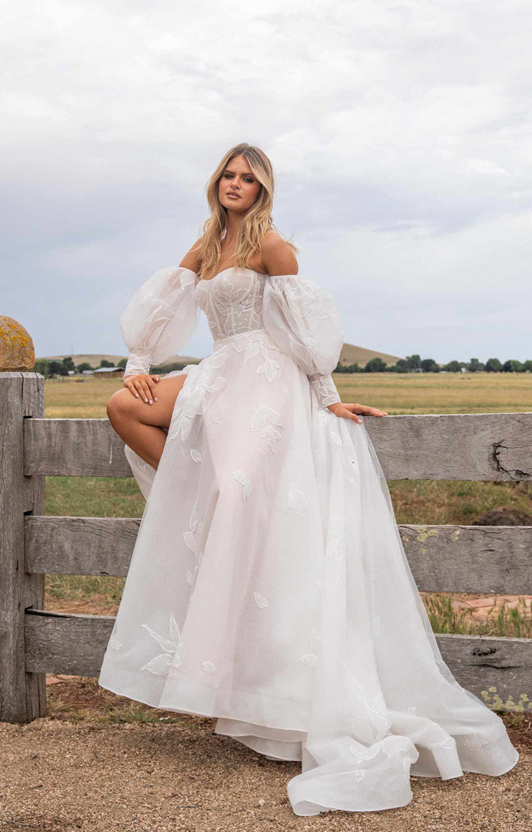 Woman in Tessa by Madi Lane, a white wedding dress sitting on a wooden fence with a field and sky in the background