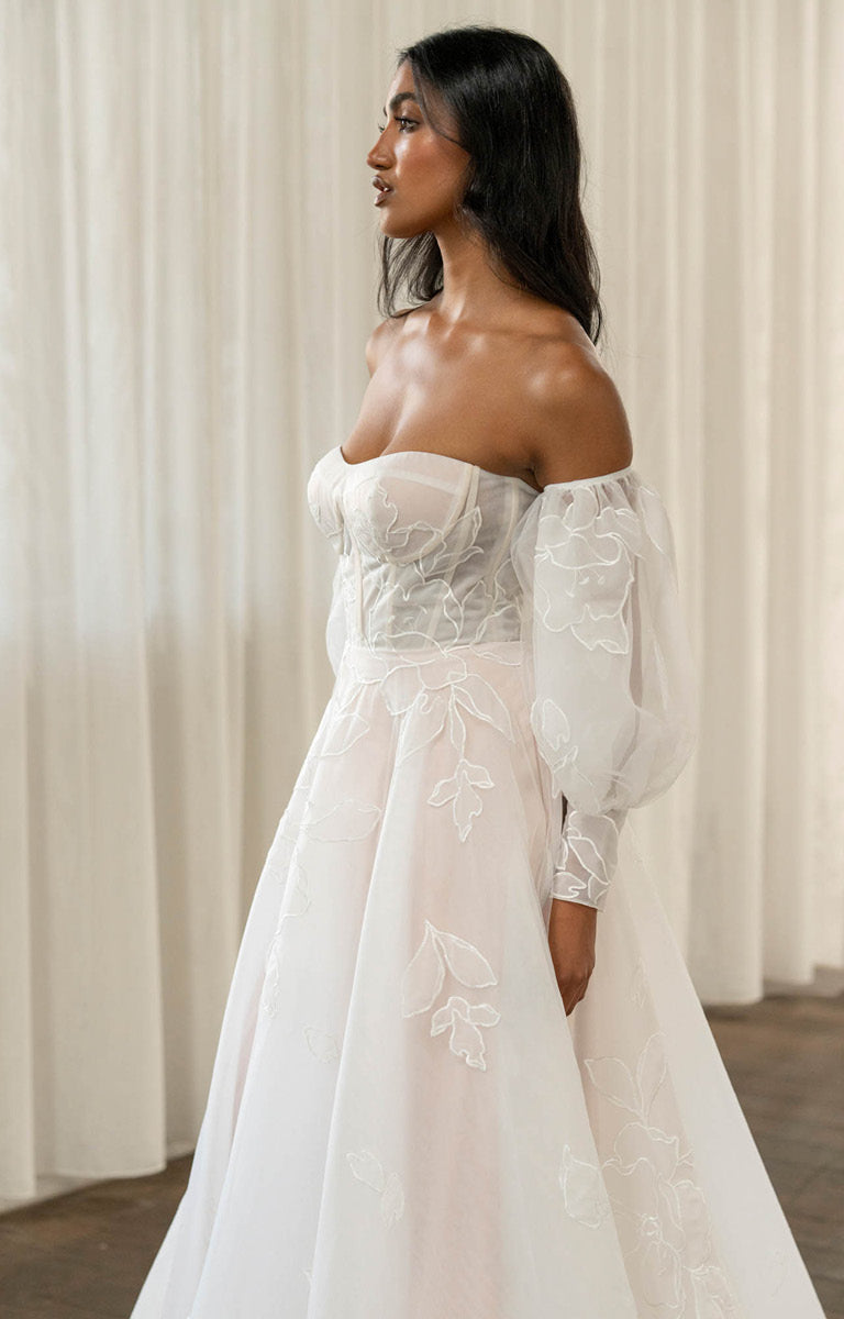Woman wearing Tessa by Madi Lane, a white lace wedding dress with long sleeves against a neutral background