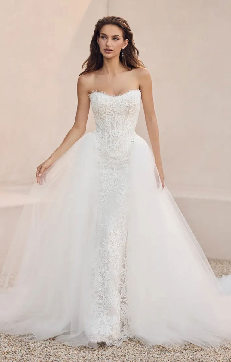 A woman modeling Mistique ST684HE by Sophia Tolli - Lace Wedding Dress with Detachable Tulle Overskirt with a fitted lace bodice, sheer lace down the front, and a voluminous tulle skirt, standing in a softly lit neutral setting.