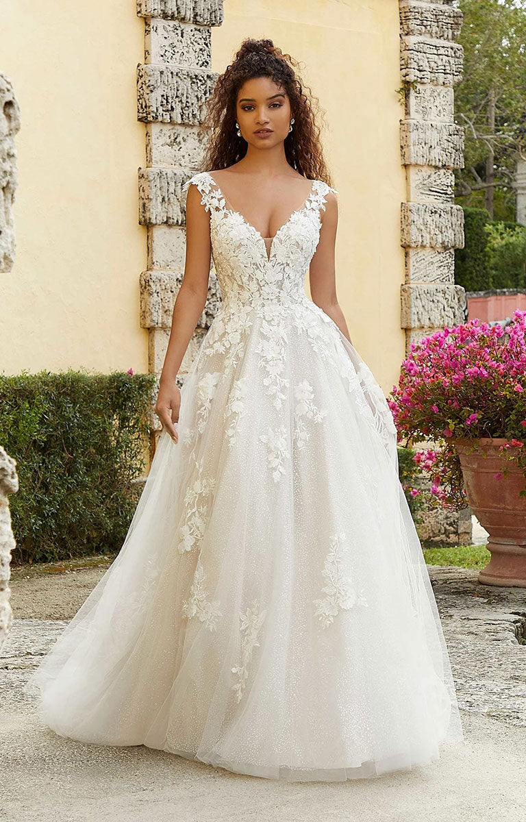 Woman in Fiorenza by Morilee - Romantic Lace and Glitter Tulle Wedding Dress with Sculpted Bodice standing in an outdoor setting with stone walls and flowers.