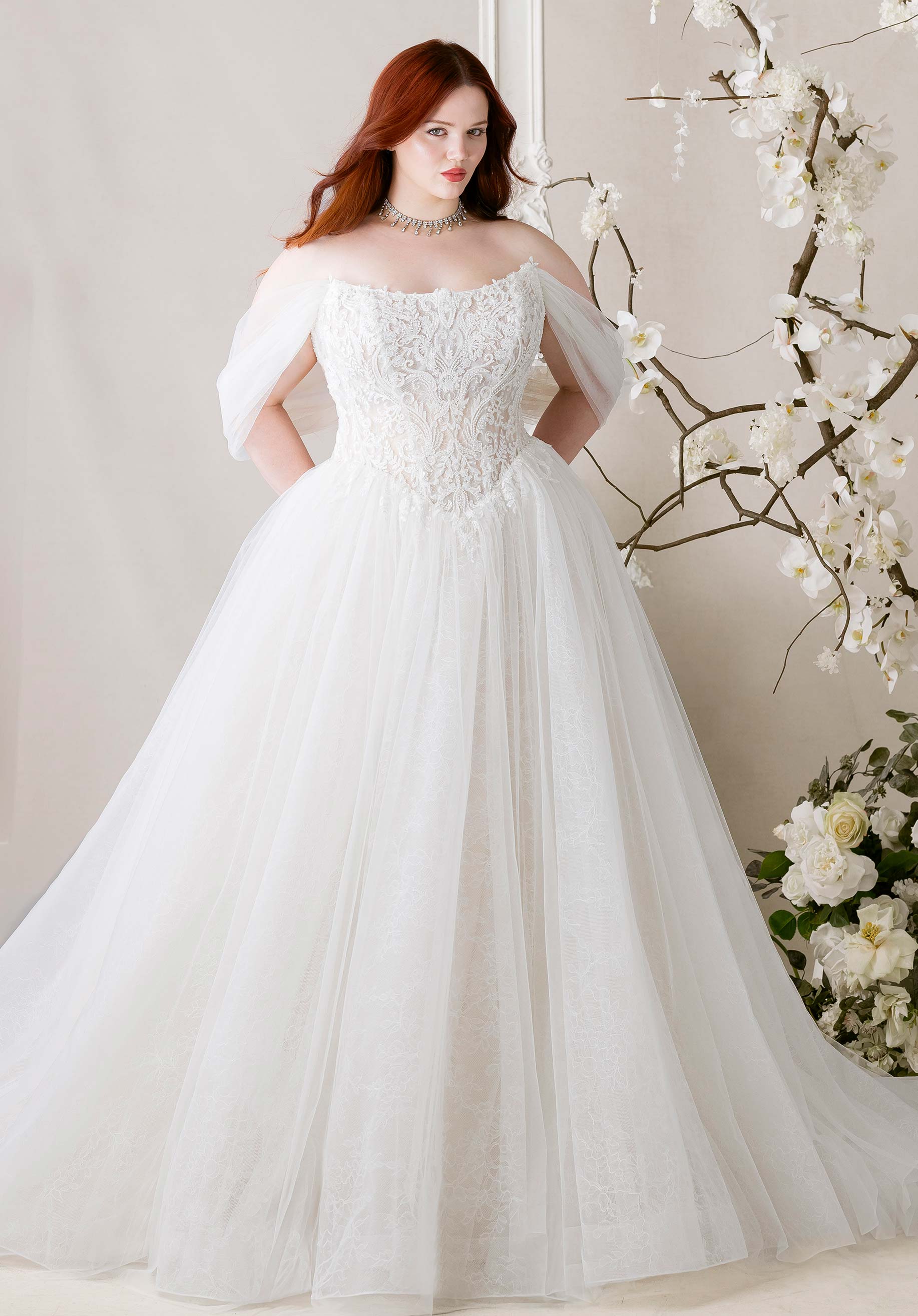 Woman wearing Meli by Morilee - Plus-Size Ball Gown with Detachable Off-the-Shoulder Sleeves with floral details in a softly lit room with flowers.