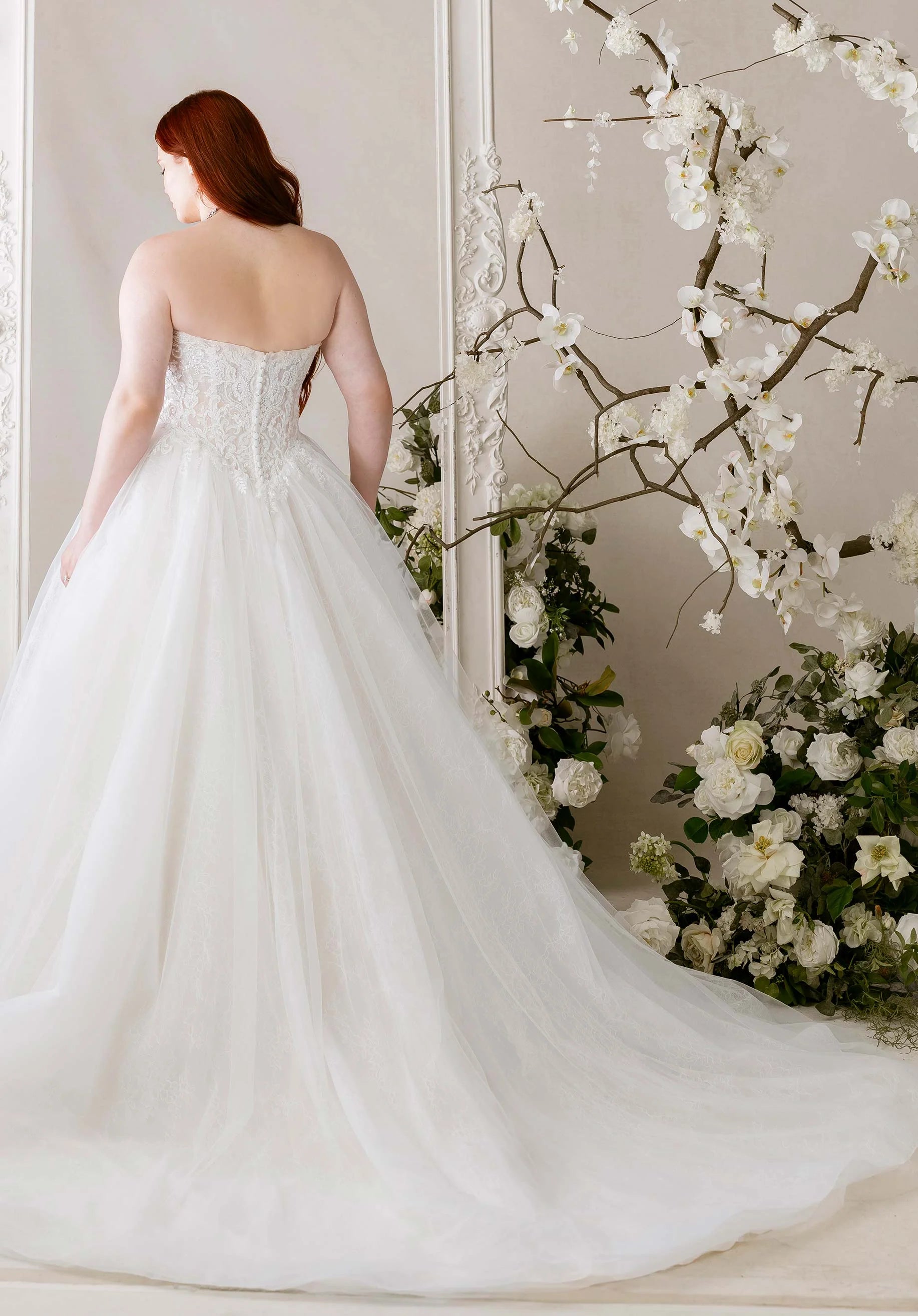 Woman in Meli by Morilee - Plus-Size Ball Gown with Detachable Off-the-Shoulder Sleeves standing in front of floral decorations