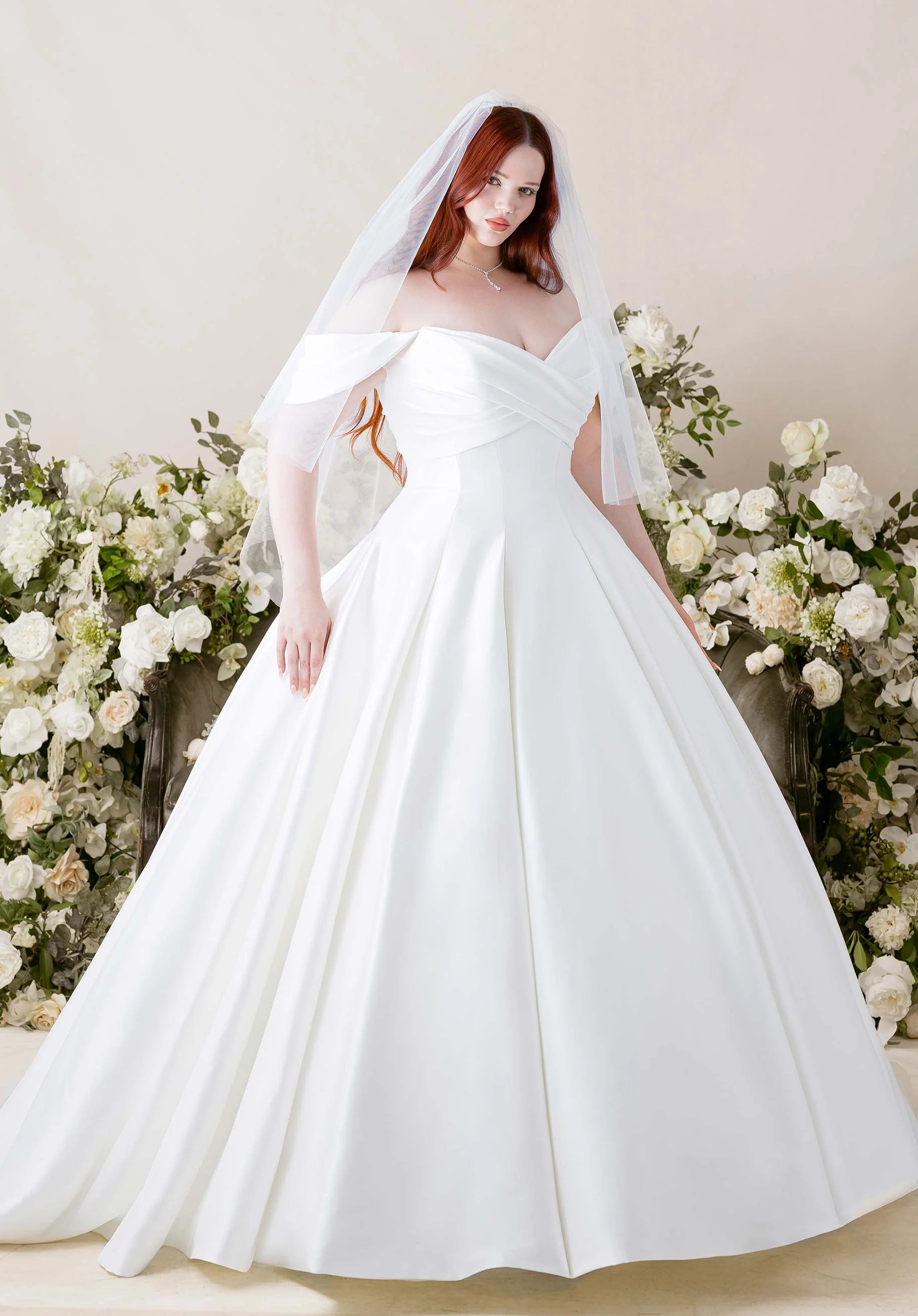 Woman in Kalina by Morilee - Ball Gown with Cat-Eye Neckline and Detachable Off-the-Shoulder Sleeves with a veil standing in front of floral decorations.