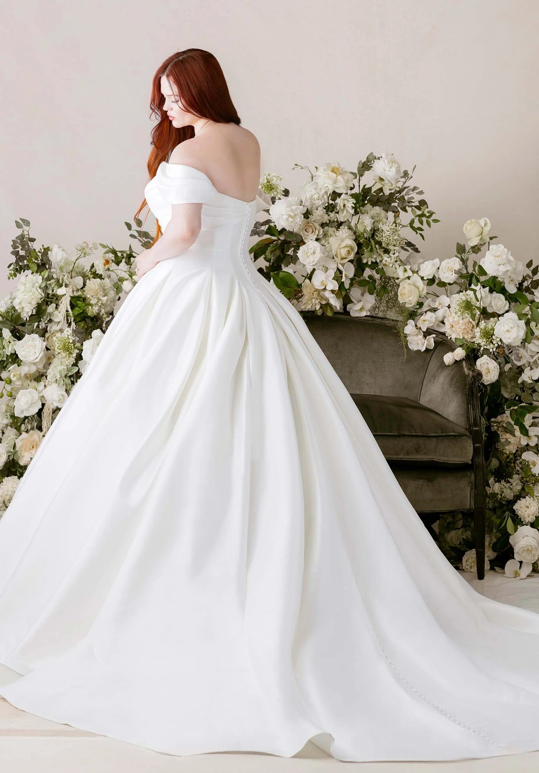 Woman in a wedding dress - Kalina by Morilee - Ball Gown with Cat-Eye Neckline and Detachable Off-the-Shoulder Sleeves standing in front of floral arrangements.