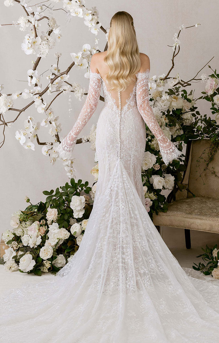 Woman in Natalie by Morilee - Fit-and-Flare Wedding Dress with Chantilly Lace, Corset Bodice and Detachable Sleeves standing in front of floral decorations.