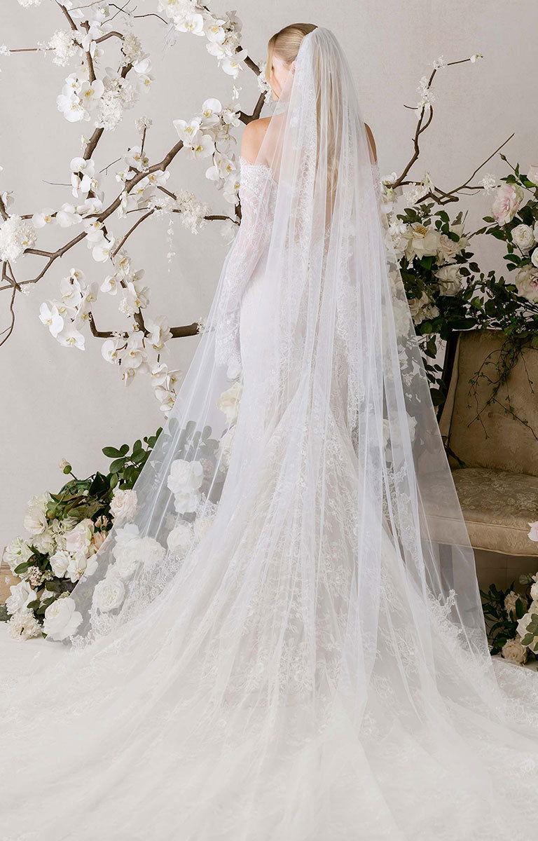 Bride in Natalie by Morilee - Fit-and-Flare Wedding Dress with Chantilly Lace, Corset Bodice and Detachable Sleeves with a long veil standing in front of floral decorations.