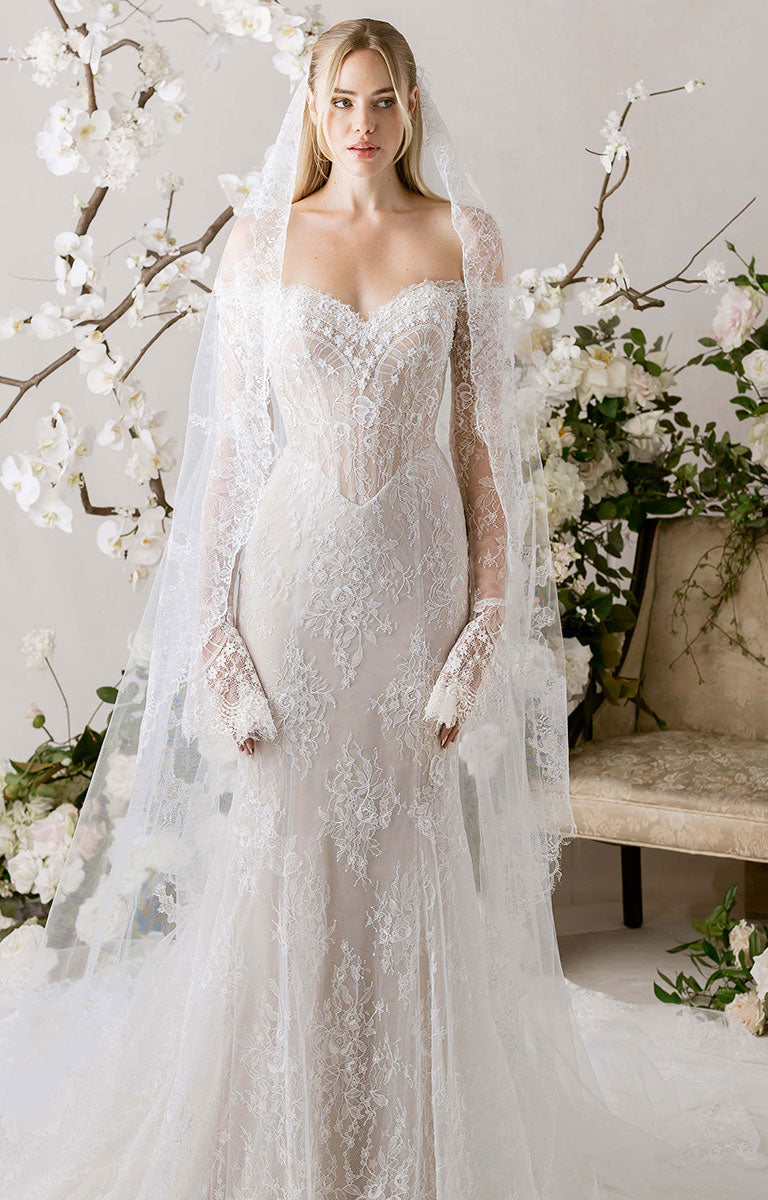 Woman in Natalie by Morilee - Fit-and-Flare Wedding Dress with Chantilly Lace, Corset Bodice and Detachable Sleeves standing in a decorated room with flowers and a chair.