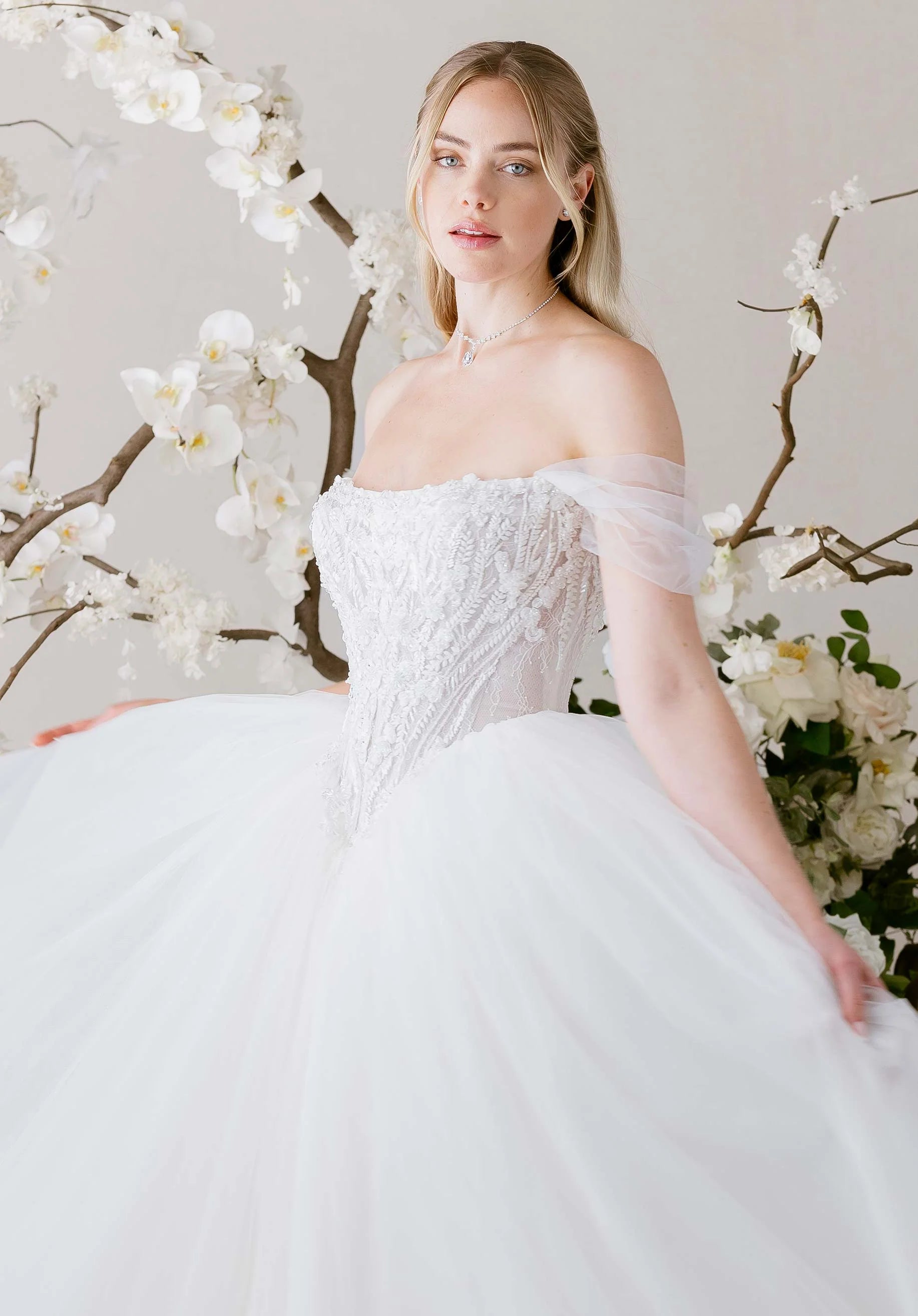 Woman in Virtuoso by Morilee - Ball Gown with Scoop Neckline and Botanical Beading standing in front of floral decorations
