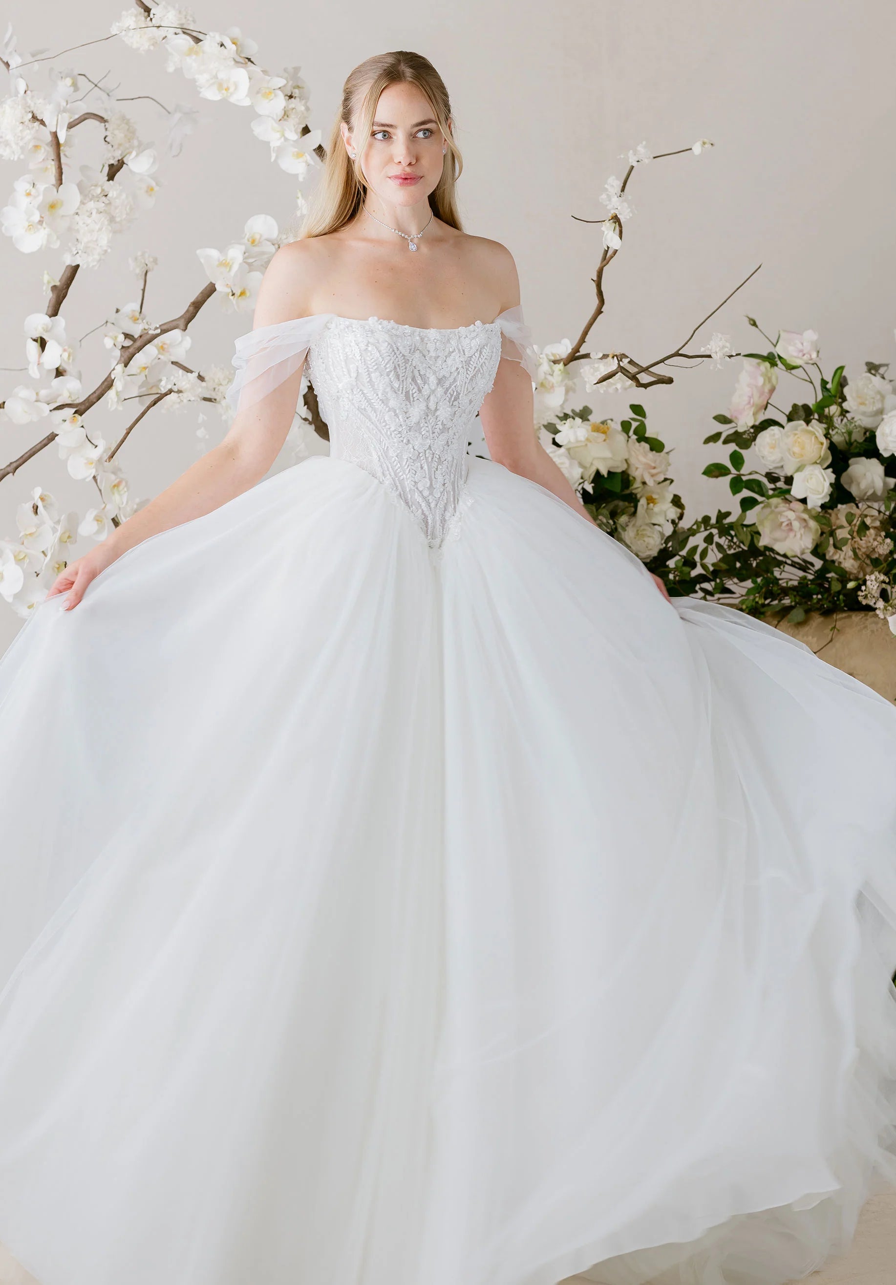 Woman in Virtuoso by Morilee - Ball Gown with Scoop Neckline and Botanical Beading standing in front of floral decorations.