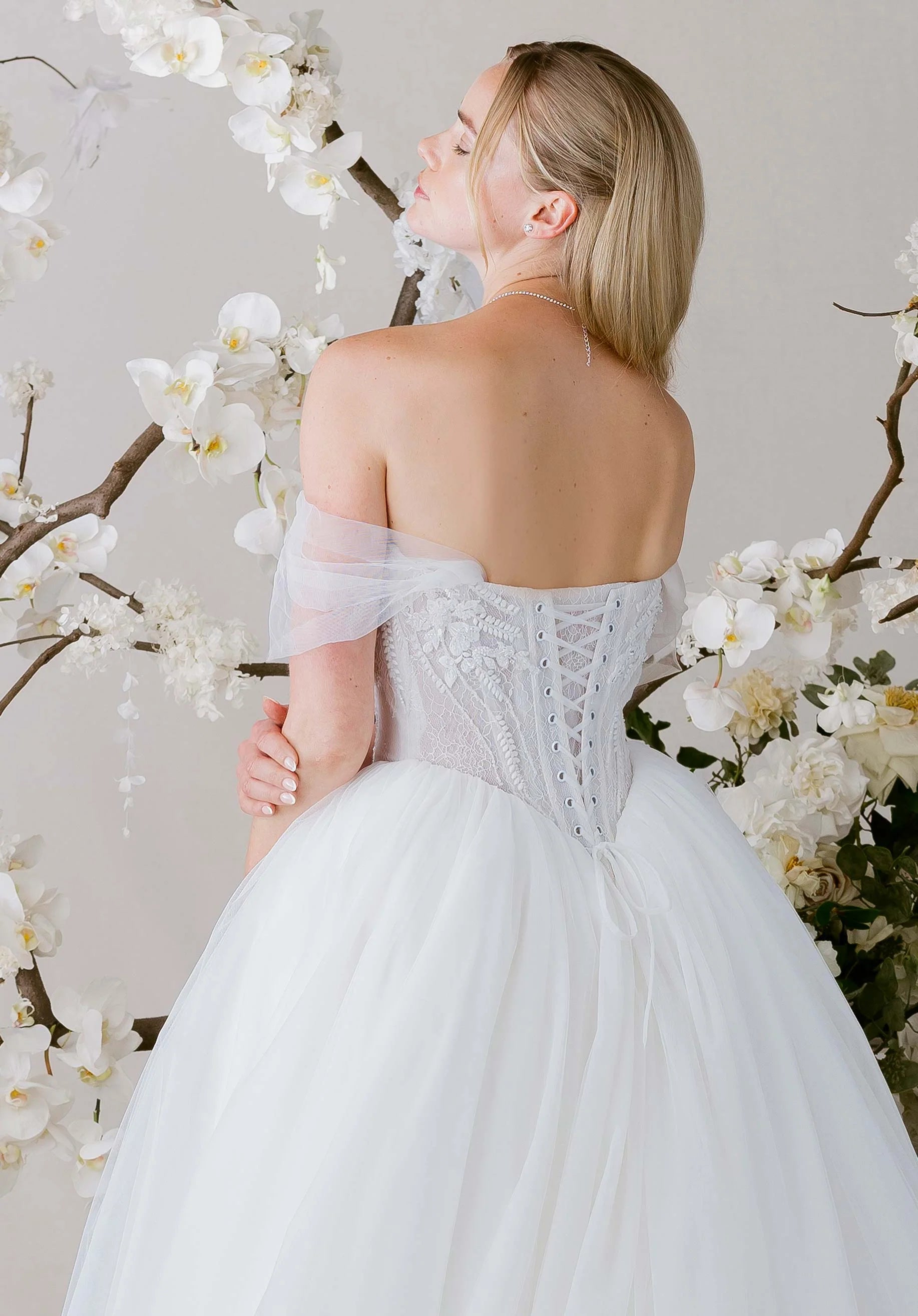 Woman in Virtuoso by Morilee - Ball Gown with Scoop Neckline and Botanical Beading standing against a floral background