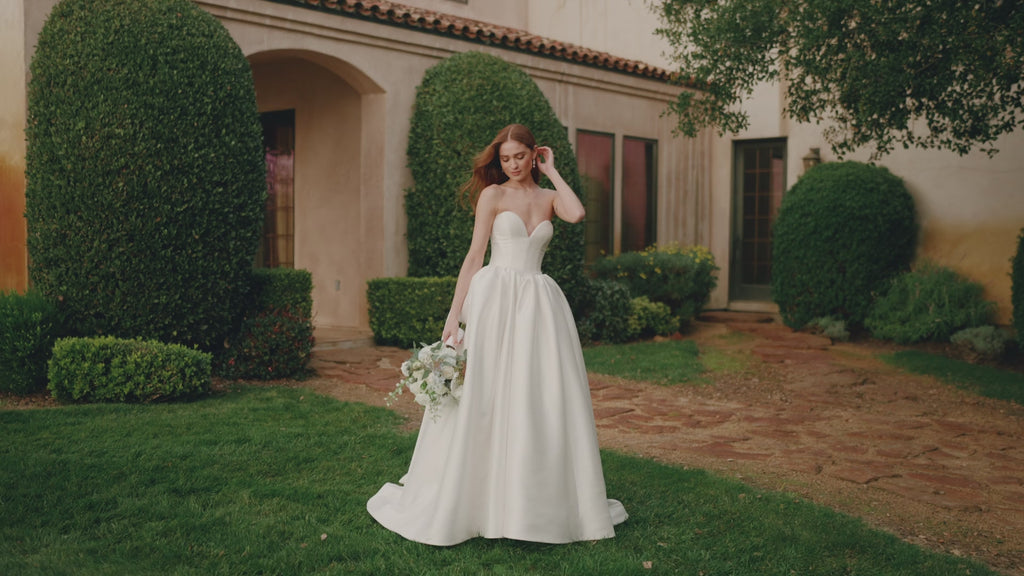 A woman wearing A1492 by Allure Bridals - Mikado Ball Gown with Basque Waist, Sweetheart Neckline and Detachable Panniers with a structured corset-style bodice and a full satin train, standing outdoors in front of a stucco house with trimmed hedges.