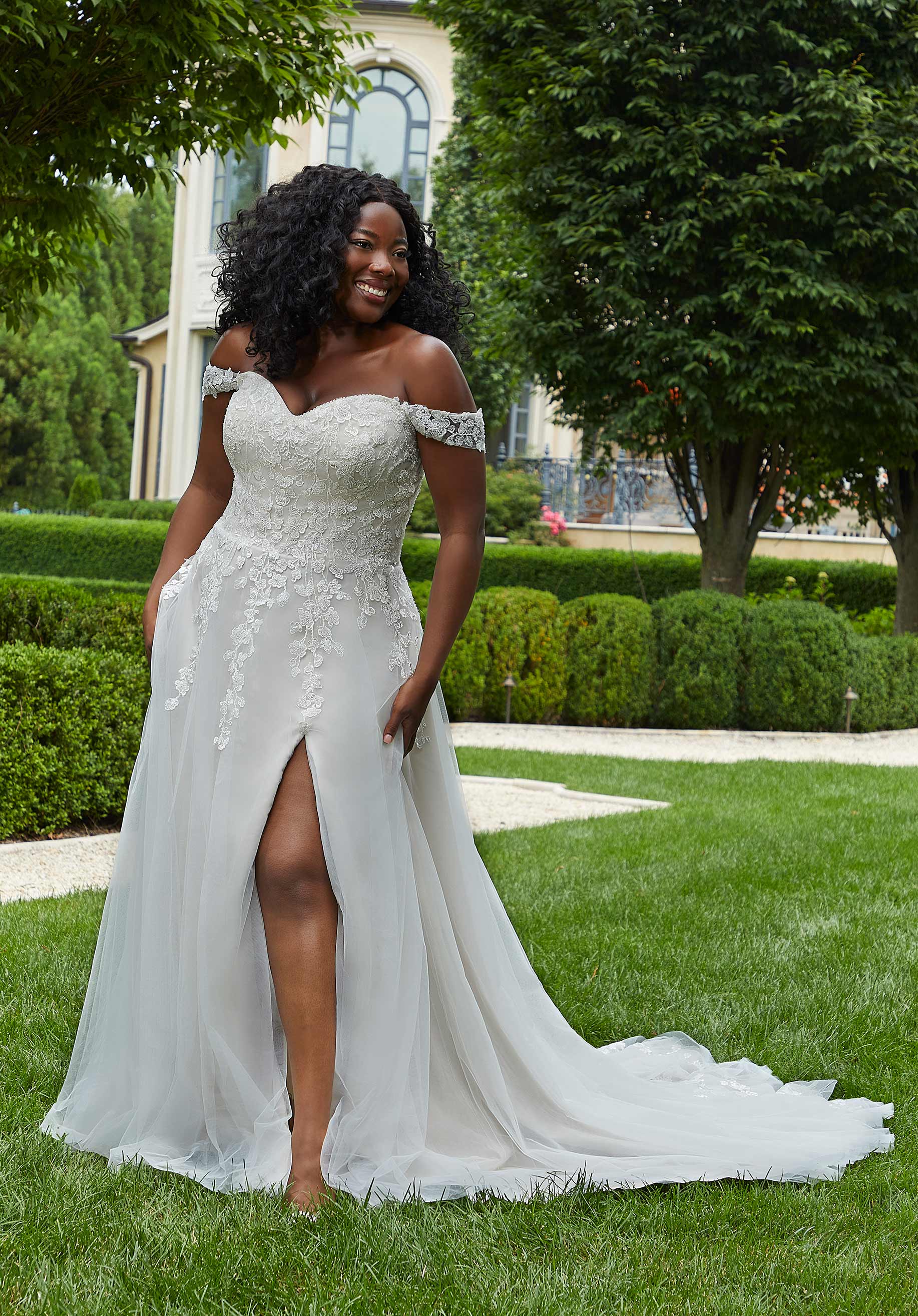 Woman in a white wedding dress standing on grass with a building and trees in the background