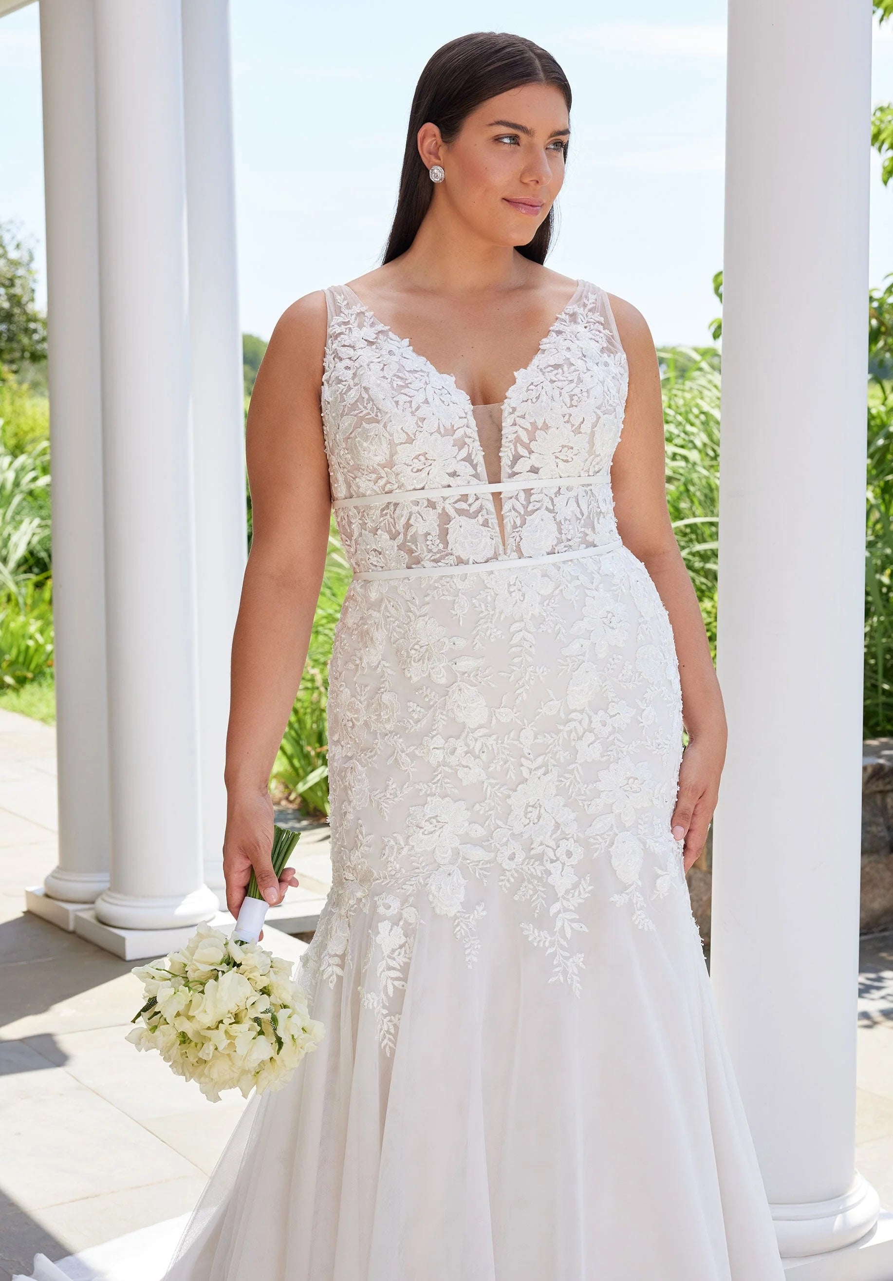 Woman in a white lace wedding dress holding a bouquet outdoors with columns in the background