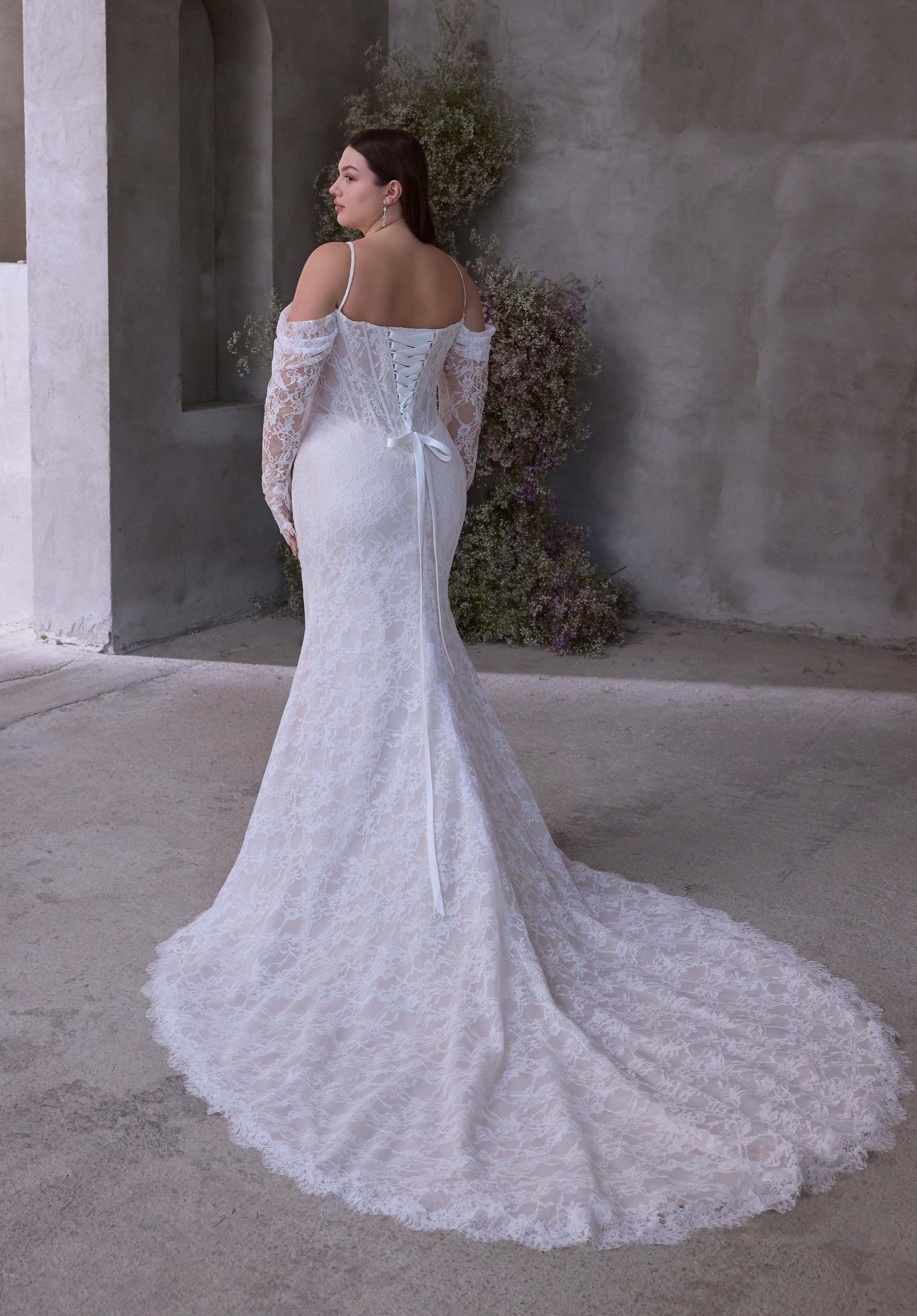 Woman in Constantia by Morilee Julietta - a white lace wedding dress standing against a neutral wall.
