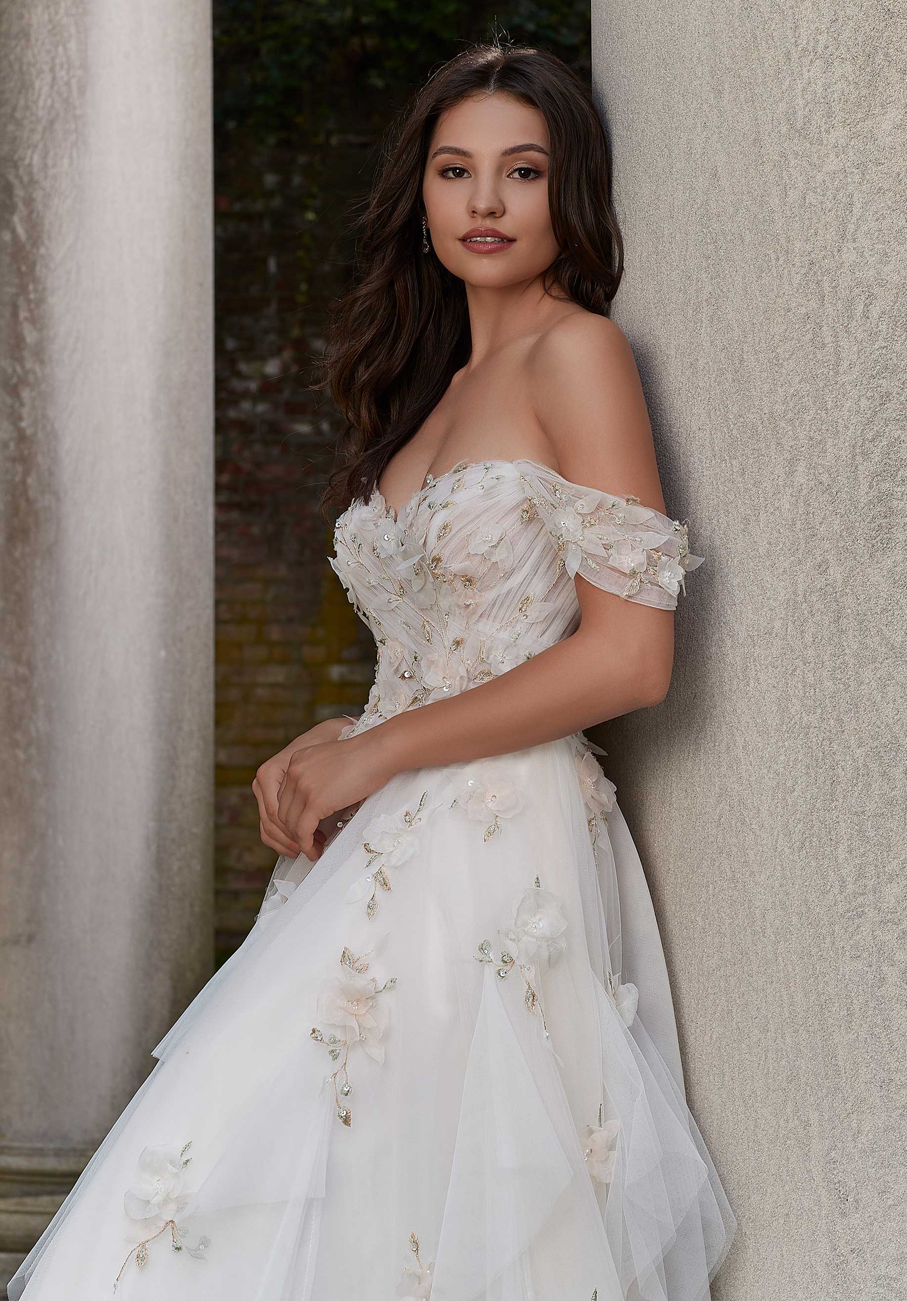 Woman wearing Petal by Morilee - a floral off-shoulder wedding dress against a textured wall.