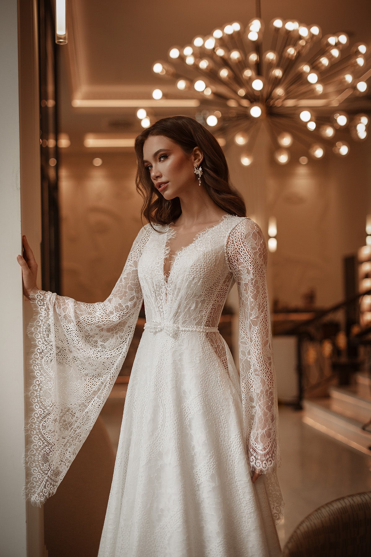 Woman in a white lace dress standing in an elegant indoor setting with chandelier.