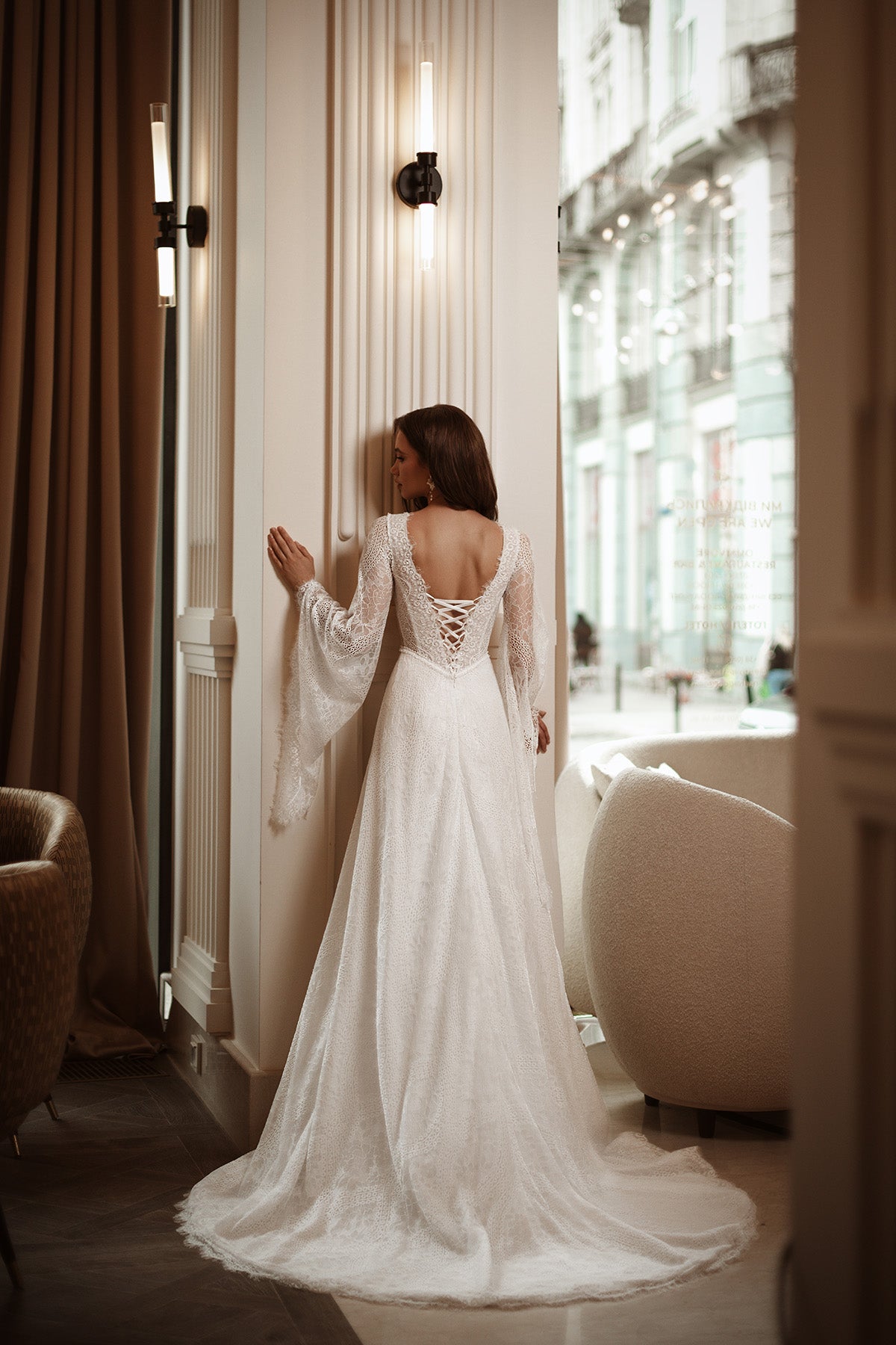 Woman in a white wedding dress standing in a luxurious room with large windows.