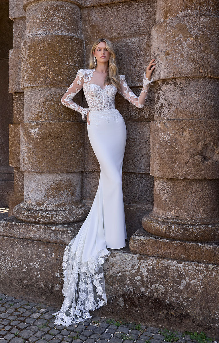 Woman in 63216 Adrianna by Veni Infantino - Fitted Wedding Dress with Botanical Lace Bodice and Illusion Sleeves standing against stone columns.