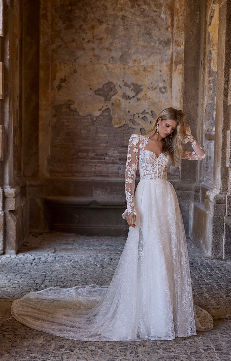 Woman in 63216 Adrianna by Veni Infantino - Fitted Wedding Dress with Botanical Lace Bodice and Illusion Sleeves standing in an old, textured stone building.