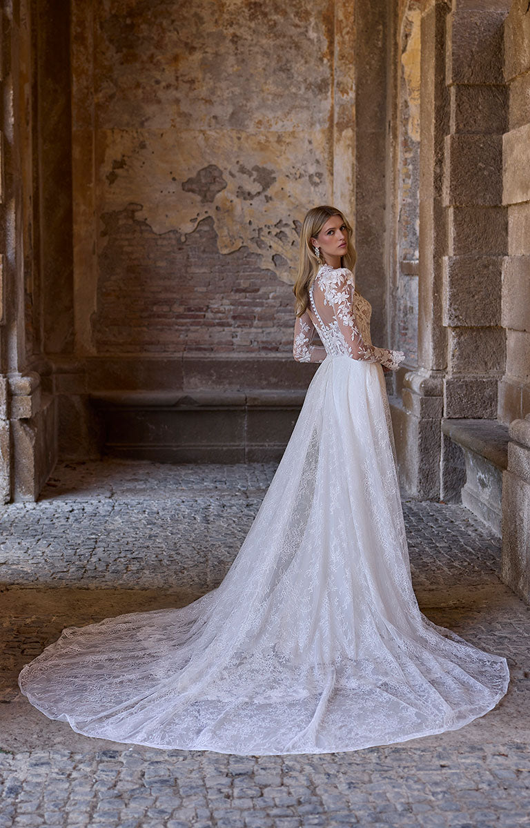 Woman in 63216 Adrianna by Veni Infantino - Fitted Wedding Dress with Botanical Lace Bodice and Illusion Sleeves standing in an ancient stone building.