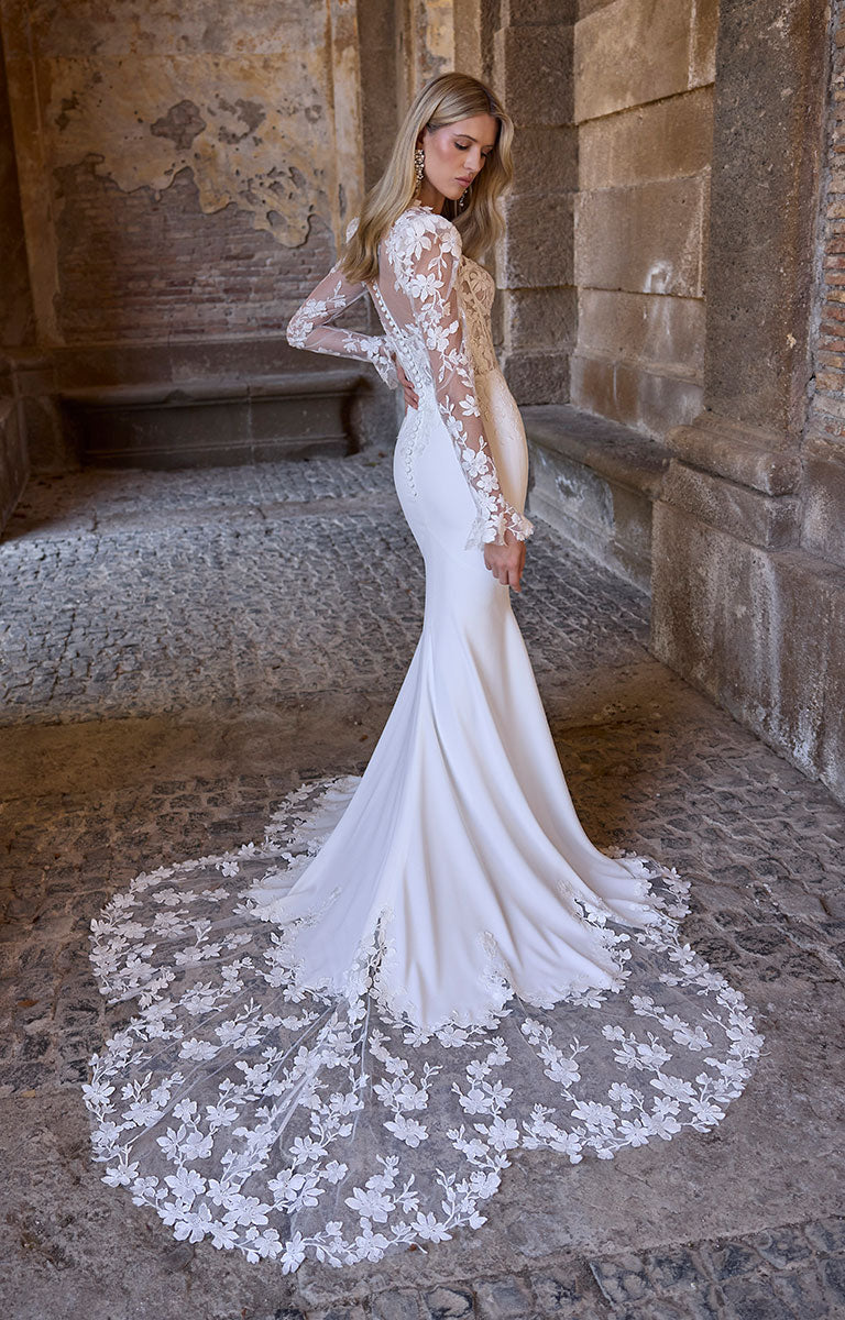 Woman in 63216 Adrianna by Veni Infantino - Fitted Wedding Dress with Botanical Lace Bodice and Illusion Sleeves with floral lace details standing in an old stone building.