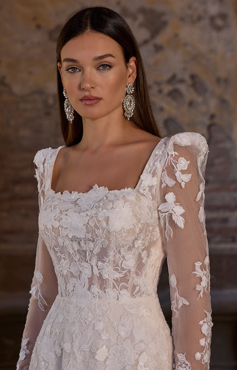 Woman wearing 63273 Apollonia by Veni Infantino - Floral Lace A-Line Wedding Dress with Square Neckline and Puff Sleeves with floral details against a blurred background