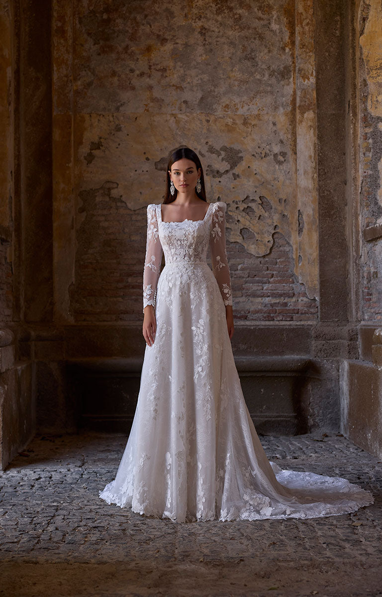 Woman in 63273 Apollonia by Veni Infantino - Floral Lace A-Line Wedding Dress with Square Neckline and Puff Sleeves standing in an old, textured wall setting