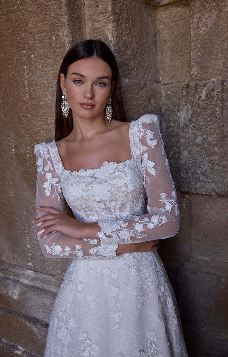 Woman wearing 63273 Apollonia by Veni Infantino - Floral Lace A-Line Wedding Dress with Square Neckline and Puff Sleeves against a stone wall.