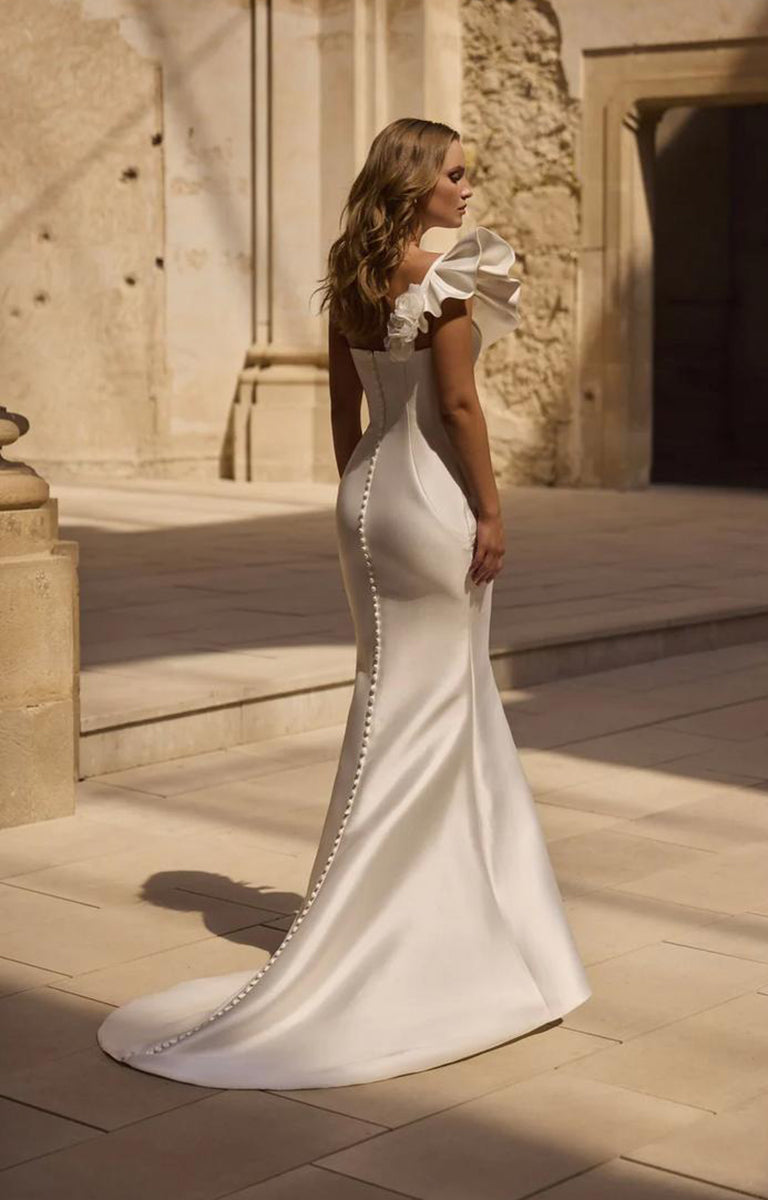Woman wearing Veni Infantino - Hatherley 63032 – Strapless Fishtail Fit and Flare Wedding Dress showing the rear of the dress with the covered buttons along the train. The photo is taken against a background with a limestone wall.