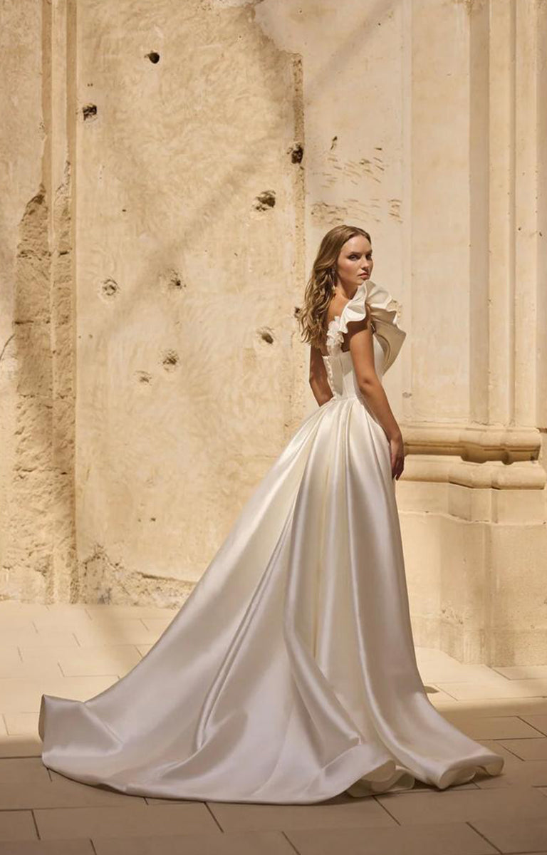 Woman wearing Veni Infantino - Hatherley 63032 – Strapless Fishtail Fit and Flare Wedding Dress against a background with a limestone wall.