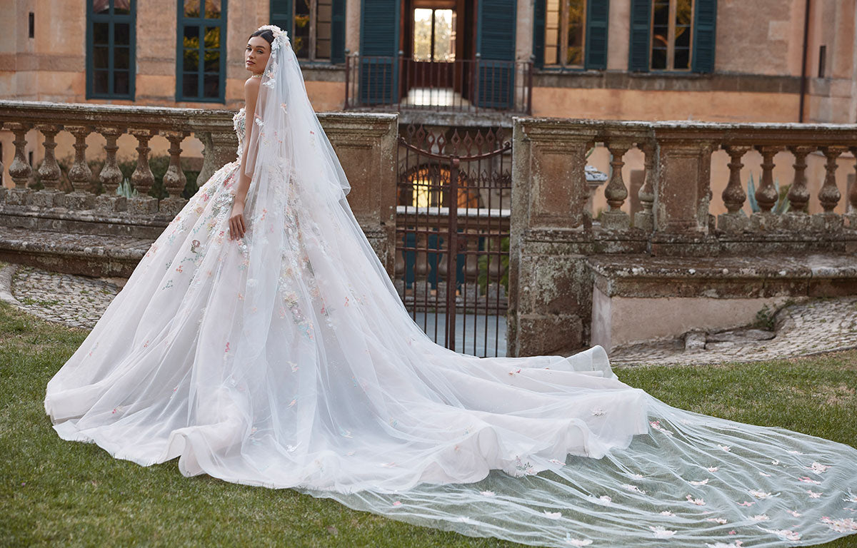 Woman in 63201A Aurora by Veni Infantino - Strapless Ball Gown Wedding Dress with 3D Floral Appliqués with a long train standing in front of an old building.
