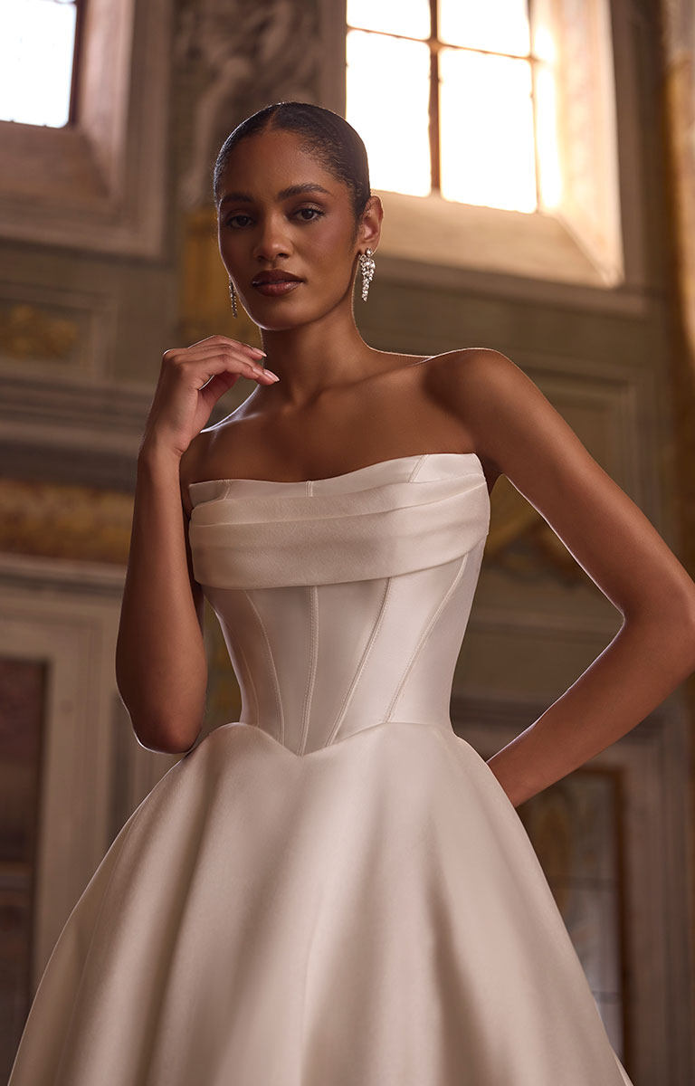 Woman wearing 63234 Avery by Veni Infantino - Strapless Mikado Ball Gown Wedding Dress with Buttoned Hem against a beautiful heritage backdrop with large leaded windows.