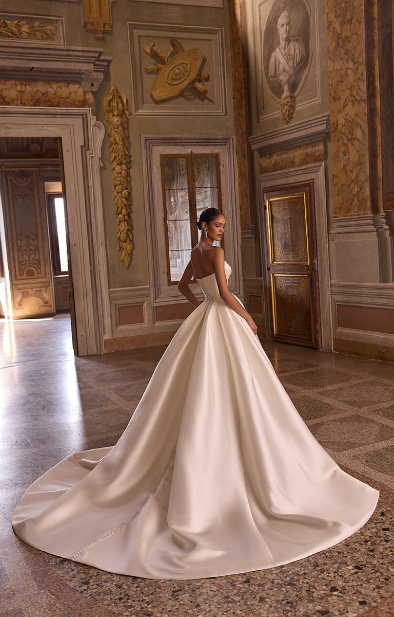Woman wearing 63234 Avery by Veni Infantino - Strapless Mikado Ball Gown Wedding Dress with Buttoned Hem against a beautiful heritage backdrop with grand artworks and murals.
