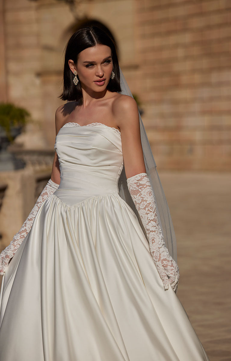 Woman in 63114 Heddie by Veni Infantino - Strapless Lace Ball Gown with Peekaboo Sweetheart Neckline with lace gloves standing against a stone wall.