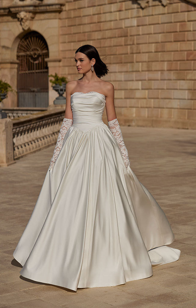 Woman in 63114 Heddie by Veni Infantino - Strapless Lace Ball Gown with Peekaboo Sweetheart Neckline standing in front of an architectural building.