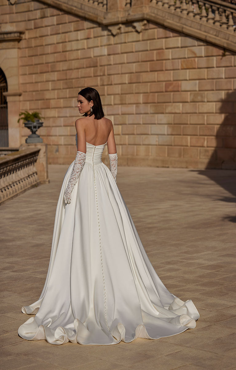 Woman in 63114 Heddie by Veni Infantino - Strapless Lace Ball Gown with Peekaboo Sweetheart Neckline standing in front of a stone building.