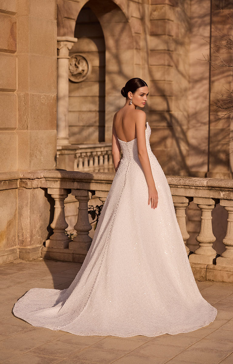 Woman in 63120 Hilani by Veni Infantino - Beaded Pearl Embellished Wedding Dress with Delicate Button Detail standing in front of a stone building.